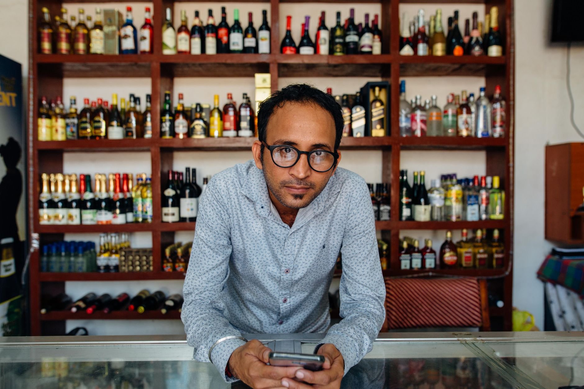 Man standing behind a counter in a liquor store, filled shelves of wine bottles behind him.