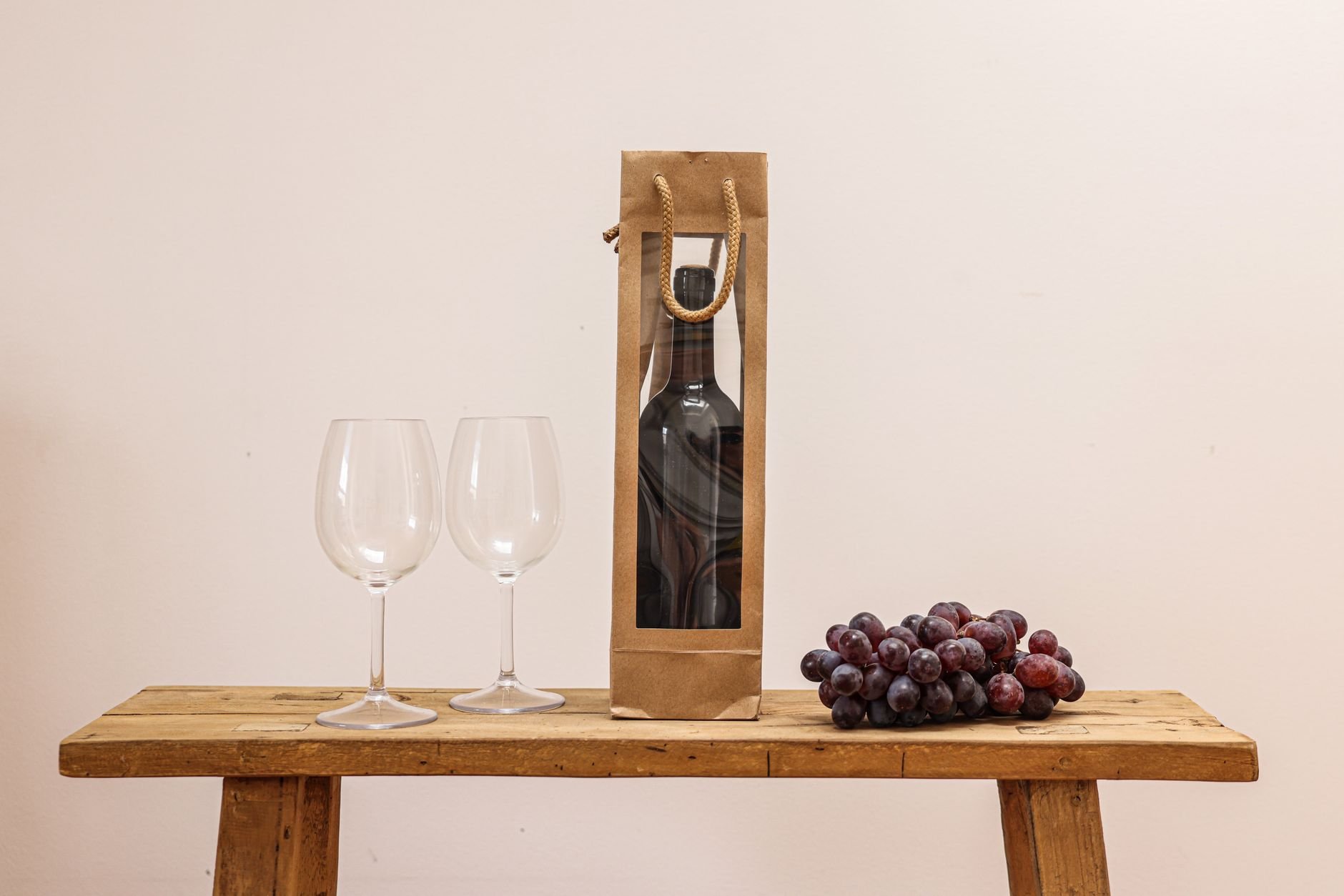 A classic still life of a wine bottle in a bag, wine glasses, and grapes on a wooden table.